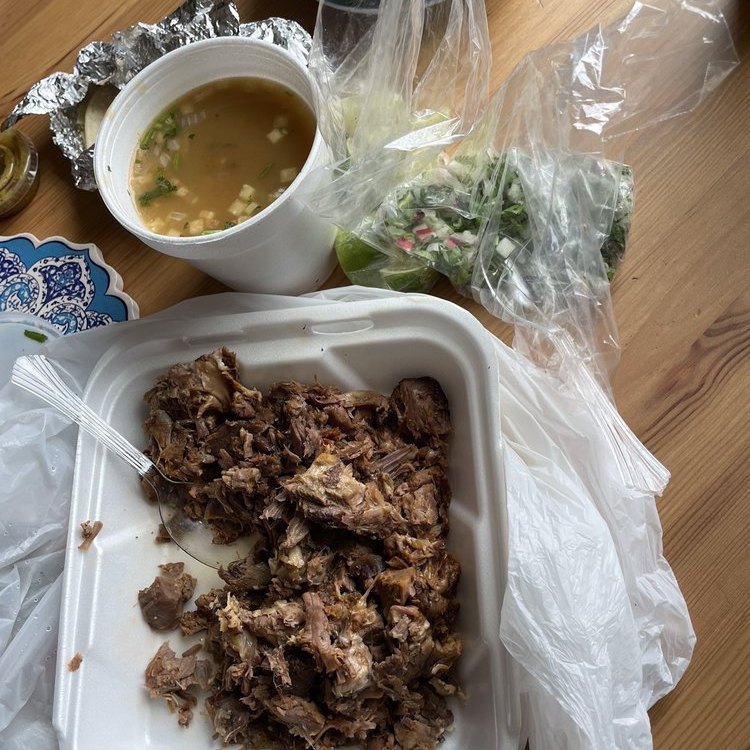 Meat in a to-go box beside a cup of broth on a wooden table